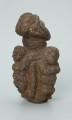 rzeźba; przedmiot obrzędowy; figura kultu zmarłych - Ujęcie z tyłu z prawej strony. Rzeźbiona w ziemistym steatycie siedząca postać ludzka o cechach kobiecych, otoczona przez trójkę mniejszych, dzieciecych postaci, skierowanych ku niej z uniesionymi rękoma.