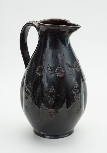 gliniany dzban zdobiony motywem roślinnym - Ujęcie z przodu skosem w prawą stronę. Dzban wykonany z gliny. Brzusiec łagodną linią przechodzi w szyjkę o pogrubionej krawędzi wylewu, z której wyodrębniony jest duży, ostro zakończony dziobek. Ucho taśmowate, wzdłużne, mocowane w połowie wysokości szyjki i w połowie wysokości brzuśca. Brzusiec zdobiony wykonanym techniką stempelkową ornamentem o motywie roślinnym. Wzór ujęty w trzy poziome pasy: dolny składa się ze schematycznie przedstawionych gron, środkowy - z pionowych elementów, na które składają się: grona, gałązka i pięciopłatkowy kwiatek, górny - z ułożonych naprzemiennie wielopłatkowych kwiatów i ulistnionych gałązek. Przy krawędzi szyjka zdobiona drobnym wytłaczanym szlaczkiem. Całość szkliwiona.