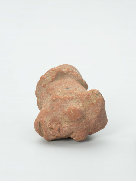 głowa - Ujęcie z góry;  Gliniana głowa postaci ludzkiej o ceglastym kolorze i prostokątnej twarzy, która opiera się na masywnej szyi. Zachowało się lewe ramię, na którym wymodelowano charakterystyczną ozdobę w kształcie koła, od którego odchodzą promienie. Figurce zaznaczono rysy twarzy: wydatne, rozchylone usta, szeroki nos o okrągłych nozdrzach oraz wypukłe, okrągłe oczy, które przypominają dwie kulki gliny. Na głowie postaci zaznaczono dwa okrągłe zgrubienia (guzki), które prawdopodobnie przedstawiają fryzurę lub nakrycie głowy. Na powierzchni widoczne są rysy, mikropęknięcia oraz przetarcia.