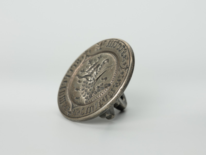 tłok pieczętny - Ujęcie z dołu skosem do lewej strony. Okrągły tłok pieczętny ze srebra z uchwytem w formie profilowanej płytki oraz wizerunkiem łba gryfa i gotyckim napisem w polu pieczęci.
