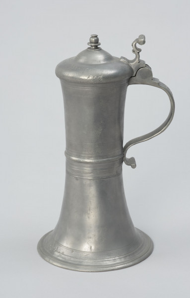 Cynowy dzban o kształcie cylindrycznym - Ujęcie z lewej strony; Wysmukły dzban o kształcie cylindrycznym z lekko wklęsłym brzuścem, z pierścieniowym zgrubieniem w centrum. Rozszerzająca się ku dołowi podstawa przechodzi w profilowaną stopę. Ucho taśmowe, silnie esowato wygięte, zakończone wałkiem. W górnej części ucha zawias podtrzymujący pokrywę z uchwytem wygiętym esowato i ozdobioną płaskorzeźbą przedstawiającą lwią głowę. Pokrywa kopulasta, dwustopniowa z profilowaną, toczoną gałką do podnoszenia na szczycie. Na brzuścu u podstawy, pośrodku i przy górnej krawędzi, a także na pokrywie delikatne profilowane i ryte pasy.