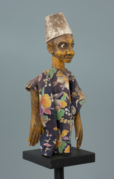 lalka teatralna: Kalhaldi (muzułmański uczony) - Ujęcie z przodu z prawej strony. Lalka teatralna przedstawiająca postać męską w nakryciu głowy w formie walca rozszerzonego u dołu, pomalowanego na biało. Szata z kwiecistego materiału, szeroka, z szerokimi rękawami. Oczy białe, z czarnymi źrenicami, drobne i wypukłe, z zaznaczonymi powiekami dolnymi oraz łukowatymi czarnymi brwiami.  Nos rozszerzony u dołu, usta zamknięte, z dolną wargą pełniejszą. Ręce przy tułowiu. Dłonie z zaznaczonymi palcami.