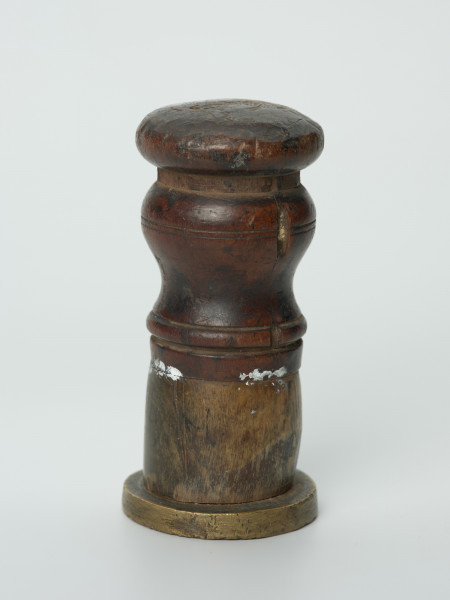 tłok pieczętny - Ujęcie w pionie pieczęcią w dół. Okragły tłok pieczętny z brązu z drewniano-rogowym uchwytem oraz rysunkiem herbu Białogardu trzymanym przez dzikich ludzi w polu pieczęci.