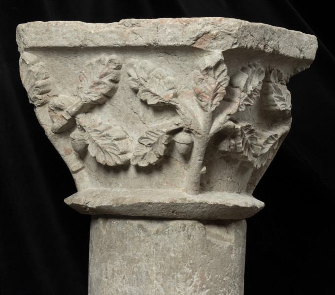 kapitel z przedstawieniem liści dębu - Ujęcie drugiego boku. Kapitel blokowo-kielichowy, rozszerzający się ku górze, wykuty z jednego kawałka kamienia. Okrągłą podstawę otacza wałek o trójkątnym profilu. Zwieńczenie w formie szerokiej czworobocznej płyty abakusa. Płaskorzeźbioną powierzchnię między wałkiem i abakusem wypełniają ustawione pionowo w narożnikach pięciolistne gałązki dębu z pojedynczym żołędziem. Mięsiste liście ukazane frontalnie w układzie niemal symetrycznym. Wyraźnie zaznaczona falista linia brzegu blaszki i wklęsły ryt unerwienia liści. Relief półplastyczny, światłocieniowy. Tło opracowane gładko. Drobne pęknięcia na powierzchni kamienia. Liczne ukruszenia zwłaszcza w partii abakusa i dolnego walka. Pod spodem centralne wgłębienie do umocowania trzpienia łączącego kapitel z trzonem.