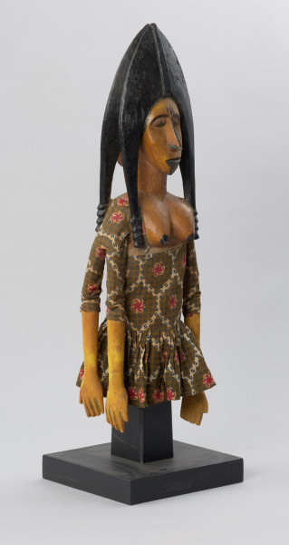 lalka teatralna: Djinè Faro (bóstwo Faro) - Ujęcie z przodu skosem w prawą stronę. Drewniana lalka przedstawiająca postać kobiecą o jasnej cerze. Twarz pociągła, nos długi, duże usta, ozdobne nakrycie głowy w kolorze czarnym. Ubrana we wzorzystą sukienkę odsłaniającą nagie piersi.