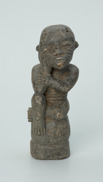 figura kultu przodków - Ujęcie z przodu. Rzeźbiona w grafitowym kamieniu postać ludzka w pozycji siedzącej - najprawdopodobniej mężczyzny. Postać siedzi na wysokiej podstawie, która swym kształtem przypomina kamień. Rzeźba ma charakterystyczne poziome nacięcia widoczne na obu policzkach. Widoczne rysy, mikropęknięcia i liczne malutkie otworki.