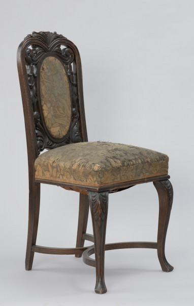 krzesło - Ujęcie z przodu, z lewej strony. Krzesło w stylu neobarokowym, oparcie i siedzisko tapicerowane. Nogi zwężające się do dołu, przednie wygięte kabriolowo, górne wybrzuszenie ozdobione płaskorzeźbionym motywem palmety, zakończenie dolne trójkątne. Tylne nogi lekko wygięte, powiązane są z przednimi krzyżową poprzeczką umieszczoną u dołu, składającą się z dwóch stykających się w środku półokrągłych pałąkowych listew. Konstrukcja zaplecka stanowi pionowe przedłużenie tylnych nóg o profilowanych krawędziach, zamykających się dwoma półkołami w kształt serca, ponad którym wyprowadzono łukowe zwieńczenie w formie mięsiście rzeźbionego, rozłożonego na boki bukietu liści. Poniżej sercowego styku listew bocznych wydłużony w pionie medalion opasany płaską listwą z wypełnieniem tapicerką. Tkanina w kwiatowy wzór w odcieniach zieleni, różu, żółcieni i ciemnego popielu. Pomiędzy owalem medalionu i listwami oparcia ażurowe wypełnienie mięsistą płaskorzeźbą o motywach roślinnych. Taką samą tkaniną obite jest siedzisko o konstrukcji sprężynowej, stosunkowo wysokie, poszerzające się do przodu, w dolnej części wykończone półwałkiem i pasmanterią, kryjącą krawędź tapicerki.