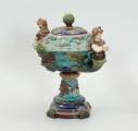 Waza - Ujęcie z przodu skosem w prawą stronę z pokrywką. Waza w kształcie smukłej amfory. Brzusiec ceramiczny, rozszerzający się ku górze, w kolorze kremowym, malowany podszkliwnie (z uzupełnieniami naszkliwnymi) we wzory kwiatowe - bukiety barwnych kwiatów w typie aster w kolorach fioletowo-czerwonym, liliowym, żółtym i białym, z gałązkami i listkami oraz drobnymi kwiatkami przypominającymi bratki o barwie fioletowo-czerwonej i liliowej. Górna część wazy wraz z uchwytami oraz stopą i część podstawy wykonane jako odlewy z mosiądzu. Mosiężna szyja wąska z otokiem perełkowym w środku ozdobiona jest pionowym, rozszerzającym się ku dołowi i ku górze rytmicznym podziałem z ornamentem geometryczno-roślinnym. Na pogrubionym skraju szyi umieszczona jest płytka miseczka w formie kwiatu o wąskich płatkach, stanowiąca wylewkę wazonu. Poniżej dolnej części szyi oprawa wierzchołka brzuśca składająca się z otoku o sercowym wycięciu zwieńczonym profilowaną palmetką oraz dwóch wydłużonych, sięgających 1/3 wysokości brzuśca, pionowych wsporników uchwytów. Te ostatnie składają się z 2 części: zewnętrzna wychodząca ze wsporników opartych na brzuścu, wyniesiona jest po niewielkim skosie do połowy wysokości szyi i na końcu wywinięta do wnętrza wolutowo, a druga - krótsza, skierowana ku dołowi łączy łagodnym łukiem wolutę z górną częścią oprawy brzuśca, kończąc się wywiniętymi na zewnątrz listkami. Uchwyty żłobkowane, zdobione motywem liści. Wazon wsparty jest na mosiężnej podstawie, składającej się z wieńca oplecionego wstęgą, na którym spoczywa zwężająca się ku górze stopa o brzegach ozdobionych perełkowaniem i pierścieniem, wspierającym koszyczek, ujmujący dolną część ceramicznego brzuśca, w formie kwiatu o ostro zakończonych, wąskich płatkach. Całość spoczywa na podstawie z kwadratowej płytki czerwonego marmuru ze ściętymi narożnikami, ujętymi mosiężnymi wspornikami ramy wspierającej płytę od dołu i opartej w narożach na czterech lwich łapach.