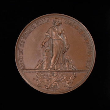 MNS/N/7342 medal nagrodowy; nagroda honorowa - Ujęcie awersu. Na awersie stojąca bogini Industria z cęgami w lewej ręce, wspierająca się prawą ręką na młocie, otoczona symbolami rzemiosła i hodowli. W odcinku scena alegoryczna: skrzydlate putto siedzące na orle, po lewej lew, po prawej delfin. Z lewej strony sygnatura. W otoku napis.