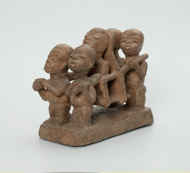 figura kultu przodków - Ujęcie przodu figury skosem z lewej strony. Rzeźbiona w brązowo-szarym kamieniu scena przedstawiająca naczelnika niesionego w lektyce przez czterech niewolników. Części twarzowe wszystkich postaci mają cechy negroidalne: wydatne, lekko rozchylone usta oraz szeroki u nasady nos. Oczy przymknięte, zamyślone. Uszy wyrzeźbiono na wysokości oczu niewolnikom, z tym wyjątkiem, że dwóm tylnym jedynie po jednym od wewnętrznej strony. Naczelnik nie ma uszu. Na twarzach postaci widoczne bruzdy na czole. Naczelnikowi zaznaczono również po dwa nacięcia na policzkach. Ręce naczelnika zgięte w łokciach, a dłonie oparte na drągach lektyki. Jego tył głowy i plecy przylegają do oparcia lektyki, natomiast zgięte w kolanach nogi ułożono jedna na drugą. Niewolników przedstawiono w pozycji stojącej. Ich powieki ozdobiono ukośnymi nacięciami. Ręce niewolników zgięte w łokciach, w dłoniach trzymają drąg od lektyki, który oparto na jednym z ich ramion. Ich nogi złączono, zaznaczono stopy. Rzeźbę umieszczono na prostokątnej podstawie, którą ozdobiono ukośnymi nacięciami.