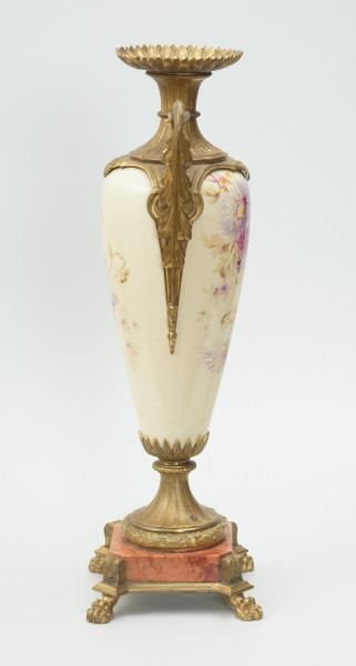 Waza - Ujęcie z prawej strony Waza dekoracyjna o bogatej, barwnej dekoracji reliefowej, w kształcie kielicha ze spłaszczoną czarą z dwoma uchwytami i przykrywą. Podstawa okrągła, profilowana z czterema prostokątnymi poszerzeniami z listkiem akantu. Trzon na styku z podstawą ozdobiony wałkiem z liści akantu, w części środkowej wypełniony reliefem z dwiema brązowymi maskami i girlandami drobnych, barwnych kwiatów. W górnej części trzonu perełkowy otok z przewężeniem na styku z czarą. Czara z szerokim brzegiem ozdobionym dekoracją geometryczną i zwężającym się do dołu brzuścem dekorowanym dwiema wypełniającymi całą wysokość czary brązowymi maskami, których zielone grzywy w formie palmety opierają się na górnej krawędzi brzegu, a brody na spodzie czary ozdobionym granatowymi 