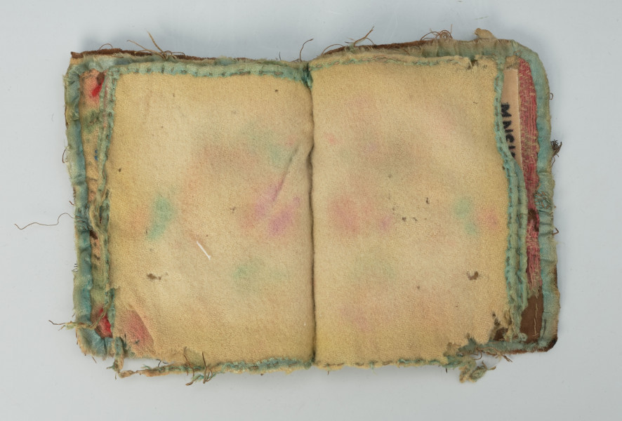 książeczka na igły z haftowanymi okładkami i kartkami z wełny - Ujęcie środka obiektu, druga strona; Przedmiot w kształcie otwartej książeczki, w której karty wykonane są z miękkiej, wełnianej tkaniny w kolorze naturalnym. Obie karty z obszytymi brzegami jasnoniebieską włóczką, bez haftu wewnątrz.