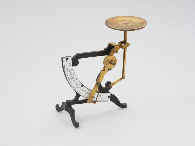 jednowahadłowa uchylna waga pocztowa - Ujęcie z tyłu prawoboczne; Jednowahadłowa uchylna waga pocztowa. Waga składa się ze stalowej skali o wartości od 0 do 350 gram, z podziałką co 5 gram, która połączona jest z trzonem wagi za pomocą stalowych blaszek znitowanych na ruchomo. Podstawa wagi składa się z dwóch elementów, z których element poprzeczny przymocowany jest do głównego elementu na ruchomo, co pozwala składać nóżkę poprzez jej przekręcenie. Dłuższe ramię podstawy zakończone jest śrubką pozwalającą na regulację wagi w opozycji pionowej. Stalowa konstrukcja łącząca główny trzon wagi z dźwignią nośną nasadzona jest na szalkę.