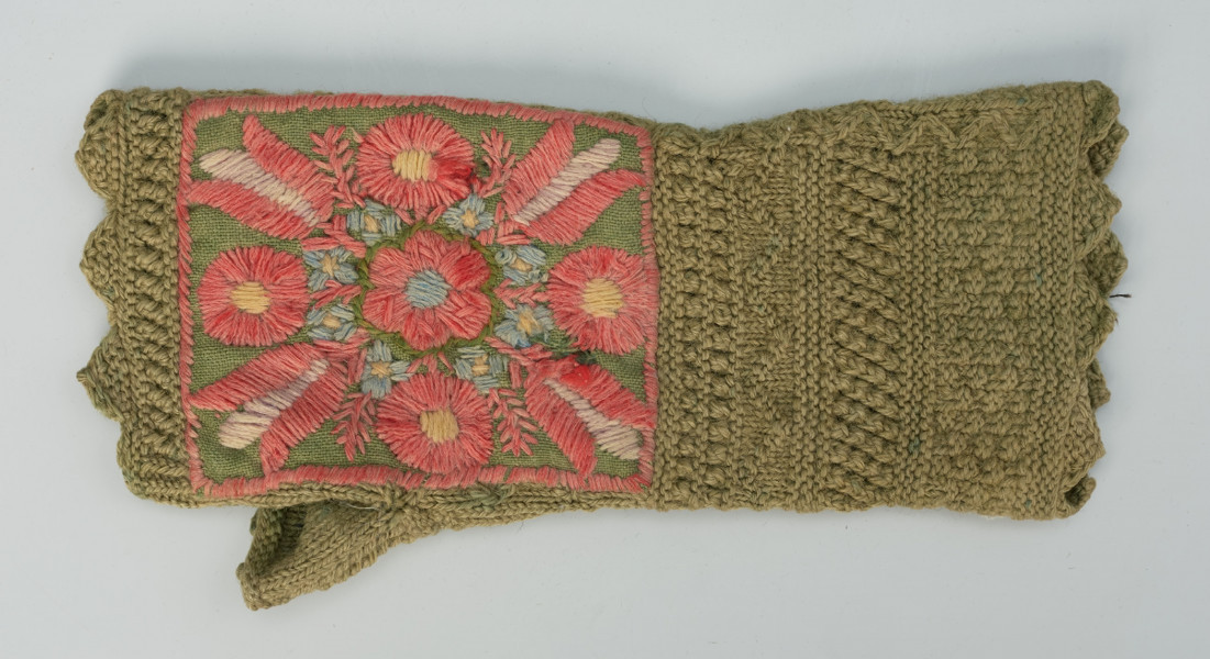 Rękawiczka bezpalcowa, z aplikacją z haftem kwiatowym - Ujęcie z przodu; Bezpalcowa rękawiczka, wykonana z zielonej wełny. Przy przegubie dłoni i przy palcach wykończona w wyraźne ząbki. Grzbiet dłoni zdobiony prostokątną aplikacją z zielonego płótna lnianego, z kompozycją wykonaną haftem płaskim. Centralnie umieszczono sześciopłatkowy czerwony kwiat z niebieskim środkiem. Wokół niego naprzemiennie wyhaftowano czerwono - żółte kwiaty. Przestrzeń pomiędzy nimi wypełniona jest drobnymi, niebieskimi kwiatkami.