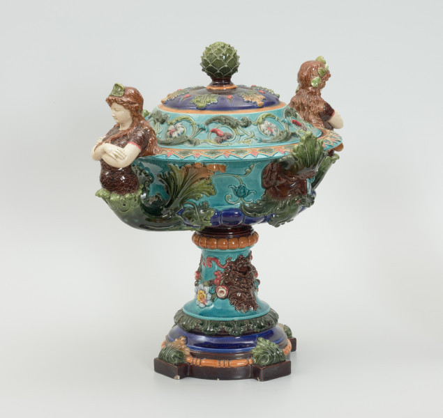 Waza - Ujęcie skosem z pokrywką. Waza w kształcie smukłej amfory. Brzusiec ceramiczny, rozszerzający się ku górze, w kolorze kremowym, malowany podszkliwnie (z uzupełnieniami naszkliwnymi) we wzory kwiatowe - bukiety barwnych kwiatów w typie aster w kolorach fioletowo-czerwonym, liliowym, żółtym i białym, z gałązkami i listkami oraz drobnymi kwiatkami przypominającymi bratki o barwie fioletowo-czerwonej i liliowej. Górna część wazy wraz z uchwytami oraz stopą i część podstawy wykonane jako odlewy z mosiądzu. Mosiężna szyja wąska z otokiem perełkowym w środku ozdobiona jest pionowym, rozszerzającym się ku dołowi i ku górze rytmicznym podziałem z ornamentem geometryczno-roślinnym. Na pogrubionym skraju szyi umieszczona jest płytka miseczka w formie kwiatu o wąskich płatkach, stanowiąca wylewkę wazonu. Poniżej dolnej części szyi oprawa wierzchołka brzuśca składająca się z otoku o sercowym wycięciu zwieńczonym profilowaną palmetką oraz dwóch wydłużonych, sięgających 1/3 wysokości brzuśca, pionowych wsporników uchwytów. Te ostatnie składają się z 2 części: zewnętrzna wychodząca ze wsporników opartych na brzuścu, wyniesiona jest po niewielkim skosie do połowy wysokości szyi i na końcu wywinięta do wnętrza wolutowo, a druga - krótsza, skierowana ku dołowi łączy łagodnym łukiem wolutę z górną częścią oprawy brzuśca, kończąc się wywiniętymi na zewnątrz listkami. Uchwyty żłobkowane, zdobione motywem liści. Wazon wsparty jest na mosiężnej podstawie, składającej się z wieńca oplecionego wstęgą, na którym spoczywa zwężająca się ku górze stopa o brzegach ozdobionych perełkowaniem i pierścieniem, wspierającym koszyczek, ujmujący dolną część ceramicznego brzuśca, w formie kwiatu o ostro zakończonych, wąskich płatkach. Całość spoczywa na podstawie z kwadratowej płytki czerwonego marmuru ze ściętymi narożnikami, ujętymi mosiężnymi wspornikami ramy wspierającej płytę od dołu i opartej w narożach na czterech lwich łapach.