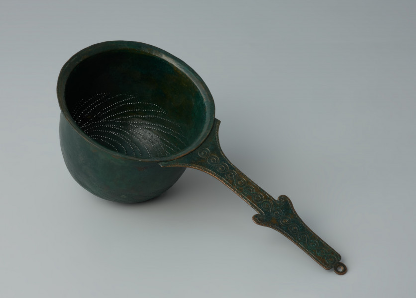 Ujęcie z góry z lewej strony. Rzymskie cedzidło z brązu z uchwytem zaopatrzonym w woluty.