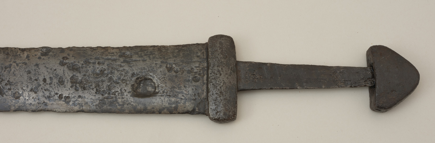broń; miecz dwusieczny - Ujecie przodu rekojeści miecza. Całkowicie zachowany. Dwusieczna głownia jest szeroka i masywna, o mniej więcej jednakowej szerokości na całej długości, przy zastawie ma przekrój sześcioboczny. Sztych jest zaokrąglony. Na obu płazach głowni widoczny wzór w postaci „jodełki” świadczący o wieloelementowej jej konstrukcji. Rdzeń został wykonany z dwóch skręconych prętów, nisko- i wysokowęglowego, tworzących jądro, z dodanymi po obu stronach nakładkami ostrzy. Trzpień miecza jest wąski i lekko zwęża się ku głowicy. Jelec jest krótki i wysoki, o kształcie widzianym od góry zbliżonym do prostokąta o lekko wypukłych krótszych bokach. Głowica jest jednolita, zbliżona do trójkąta. Jelec i głowica są lite, wykute każde z pojedynczego kawałka metalu.