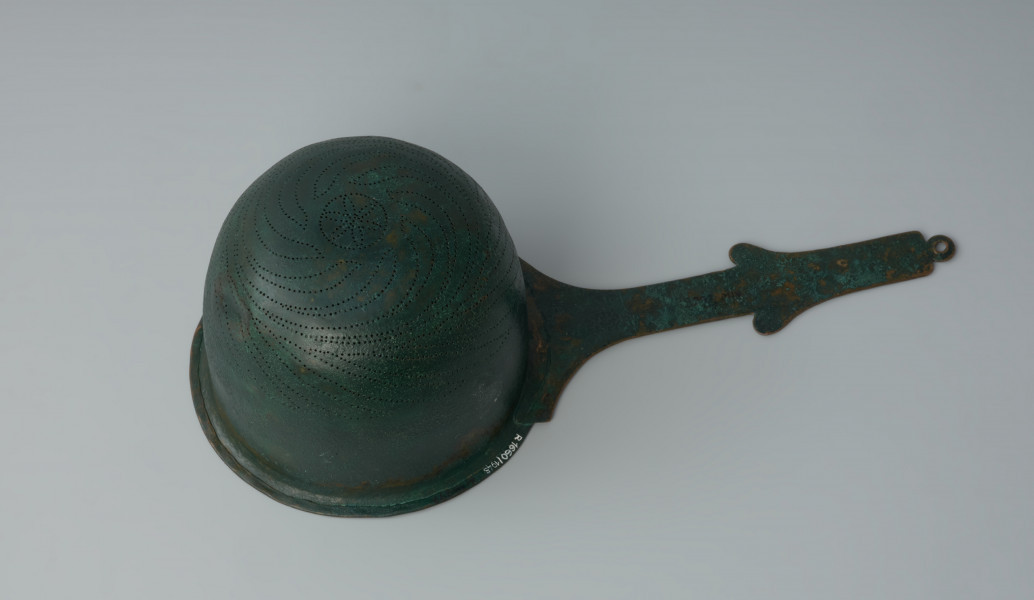 Ujęcie z dołu z prawej strony. Rzymskie cedzidło z brązu z uchwytem zaopatrzonym w woluty.