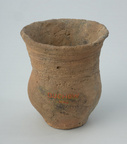 Naczynie gliniane - Ujęcie z tyłu; Puchar o kształcie esowatym, zdobiony na szyjce dookolnymi pasmami odcisków sznura, złożonymi z 4 grup odcisków zwielokrotnionych linii, podkreślonym od dołu dookolną linią drobnych nakłuć. Powierzchnia nierówna, niestarannie wyrównywana, ze śladami po wypadających w trakcie wypału dodatkach w masie garncarskiej. Nad dnem wywiercony wtórnie otworek. Naczynie rekonstruowane i uzupełnione barwionym gipsem.