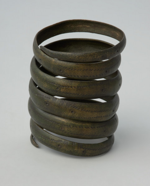 naramiennik spiralny typu kujawskiego - Ujęcie w pionie. Sześciozwojowy naramiennik wykonany z wąskiej brązowej taśmy. Powierzchnię taśmy zdobi ornament oczkowy połączony z ukośnymi nacięciami i poprzecznymi żłobkami. Starannie opracowane zakończenia naramiennika są uformowane w szpic.