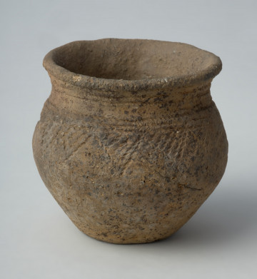 Naczynie gliniane - Ujęcie z przodu; Puchar o kształcie esowatym z krotką szyjką, zdobiony na szyjce dookolnymi pasmem odcisków sznura złożonym z 3 linii oraz poniżej nasady brzuśca podobnym pasmem, z którego zwieszają się ukośnie wypełnione trójkąty.