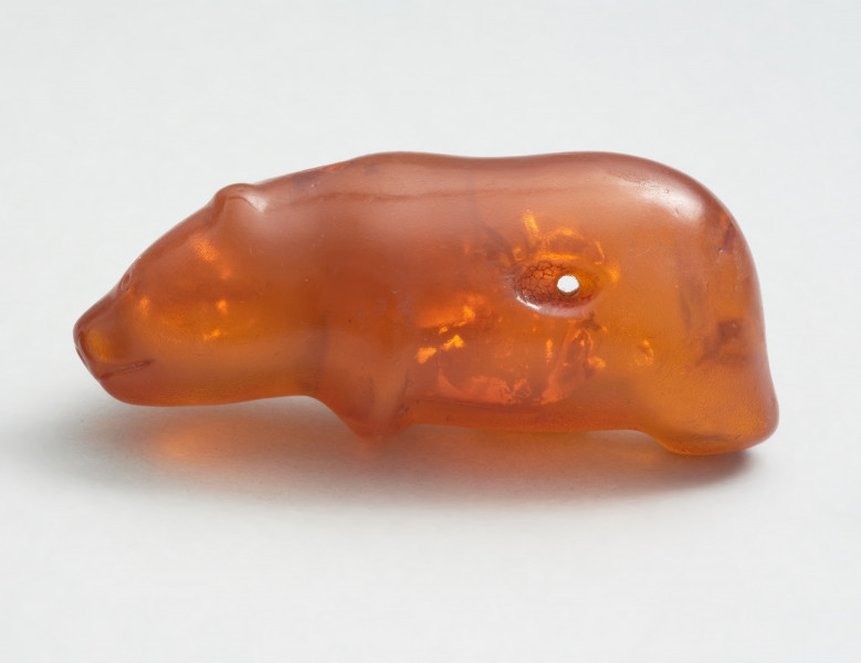 bursztynowa figurka niedźwiedzia ze Słupska - Ujęcie z lewej strony; Bursztynowa figurka niedźwiedzia. Figurka ma schematycznie przedstawiony tułów i nogi oraz realistycznie oddaną głowę. W momencie znalezienia powierzchnia figurki była matowa i spatynowana, jednak krótko po odkryciu przeprowadzono konserwację zabytku. Także ostro zarysowane oczy, usta i nozdrza są wynikiem prac konserwatorskich. Prawdopodobnie oryginalnie miały bardziej schematyczną formę. Figurka posiada dziurę w tułowiu, która jest efektem formowania się bryłki bursztynu wokół gałązki, która z czasem uległa dekompozycji.