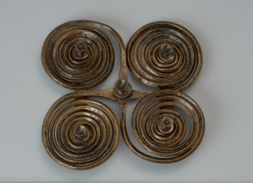 Zapinka krzyżowa - Ujęcie z góry. Zapinka krzyżowa wykonana jest z dwóch brązowych prętów o rombowatym przekroju, z których uformowano po parze spiralnych tarczek, nadając im ósemkowate kształty. Czworokątny kształt uzyskano składajac na krzyż obydwa elementy i spajając je od spodu dwoma wąskimi blaszkami, z których jedna zachowana jest w połowie. Fibulę udekorowano pięcioma ozdobnymi guzkami.