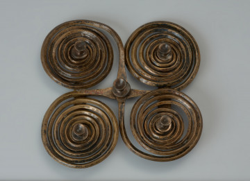 Zapinka krzyżowa - Ujęcie z góry. Zapinka krzyżowa wykonana jest z dwóch brązowych prętów o rombowatym przekroju, z których uformowano po parze spiralnych tarczek, nadając im ósemkowate kształty. Czworokątny kształt uzyskano składajac na krzyż obydwa elementy i spajając je od spodu dwoma wąskimi blaszkami, z których jedna zachowana jest w połowie. Fibulę udekorowano pięcioma ozdobnymi guzkami.