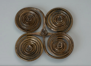 Zapinka krzyżowa - Ujęcie z góry. Zapinka krzyżowa wykonana jest z dwóch brązowych prętów o rombowatym przekroju, z których uformowano po parze spiralnych tarczek, nadając im ósemkowate kształty. Czworokątny kształt uzyskano składajac na krzyż obydwa elementy i spajając je od spodu dwoma wąskimi blaszkami, z których jedna zachowana jest w połowie. Fibulę udekorowano pięcioma ozdobnymi guzkami.