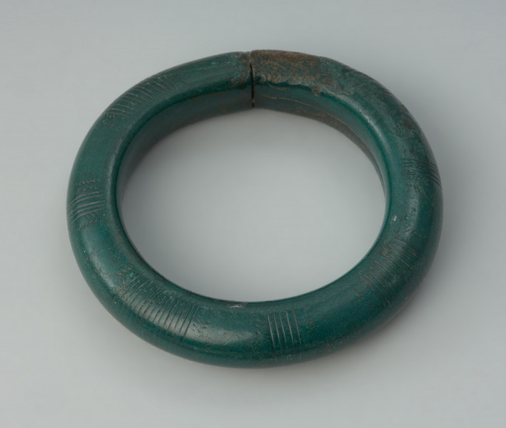 Bransoleta obręczowa - Ujęcie z góry całej bransolety. Masywna bransoleta wykonana z grubego brązowego pręta o przekroju kolistym. Końce obręczy są ścięte prosto i ściśle do siebie przylegaja. Powierzchnię zabytku pokrywa delikatnie wyryte zdobienie złożone z grup poprzecznych kresek, połączonych z motywem jodełki. Bransoletę pokrywa ciemnozielona patyna.