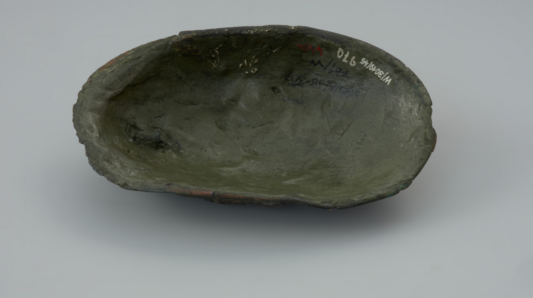 zapinka miseczkowata - Ujęcie spodu. Zapinka ma kształt wydłużonego owalu. Przedmiot jest częściowo uszkodzony bez zachowanej szpili. Powierzchnia fibuli jest bogato ornamentowana motywami zoomorficznymi. Na detal zdobniczy składa się osiem pól wyznaczonych przez krzyżujące się wstęgi. W punktach ich przecięcia znajdują się ślady po siedmiu wypukłych guzkach. Na sześciu z ośmiu pól znajdują się motywy zwierzęce bliskie skandynawskiej sztuce w stylu Borre. Na trzech z czterech zachowanych pól zlokalizowanych w narożnikach fibuli występują zwierzęta chwytające swoimi szponami ciała innych stworzeń. Dwa kolejne naprzeciwległe pola, usytuowane w środkowej części odpowiadają dwóm łukowato wygiętym w biodrach zwierzętom - być może są to konie z głowami skierowanymi w swoją stronę. Pozostałe dwa pola zawierają przedstawienia zwierzęcych masek z połączonymi ze sobą zaciśniętymi łapami.