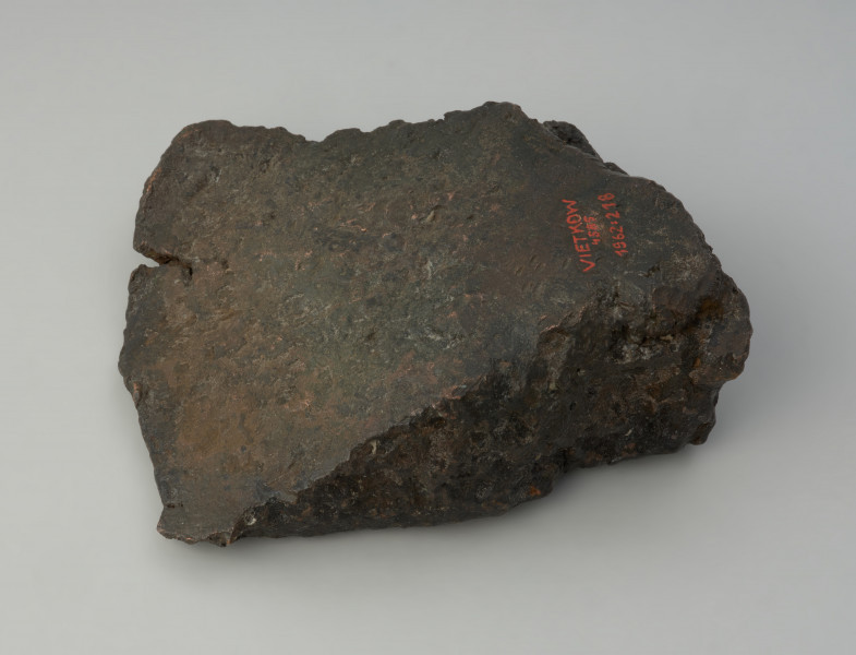 bryła metalu - Ujęcie w poziomie tyłu skosem w lewą stronę. Fragment placka odlewniczego o nieregularnym, czworobocznym kształcie z zachowanym jednym z pierwotnych boków o ostrej krawędzi. W przekroju poprzecznym trójkatny.