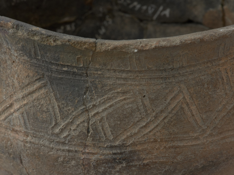 naczynie gliniane - Ujęcie fragmentu górnej części naczynia z widocznym wzorem. Zdobienie w postaci krzyżujących się skośnych linii wpasowanych w podwójne, dookolne linie ryte.