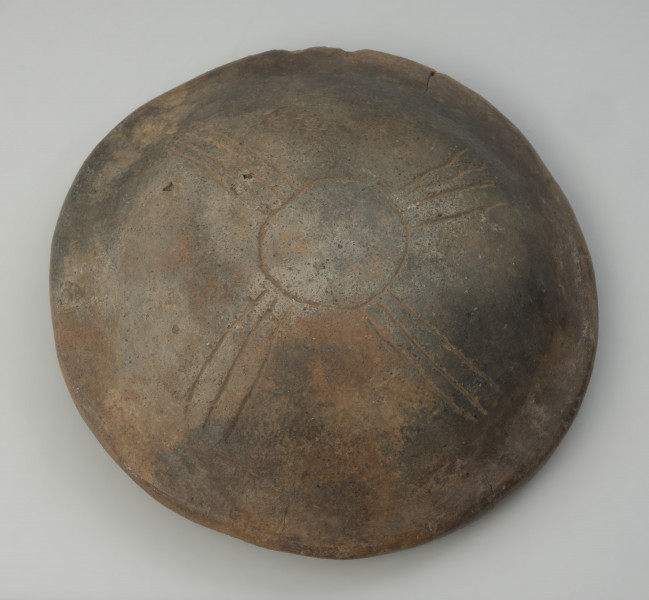 naczynie sepulklarne, urna twarzowa - Ujęcie spodu. Urna twarzowa z zaznaczonymi cechami twarzy ludzkiej, uszami ozdobionymi brązowymi kolczykami, ornamnetem rytym sugerującym naszyjnik oraz z pokrywką w kształcie kapelusza.