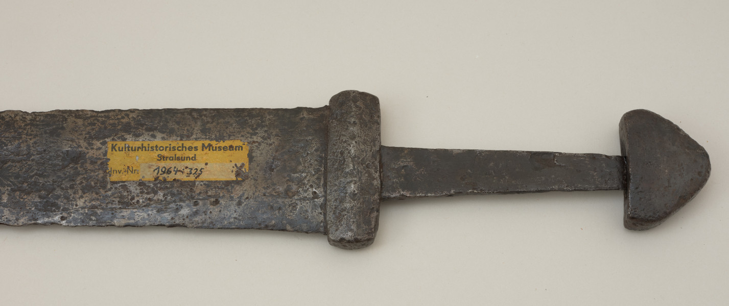 broń; miecz dwusieczny - Ujęcie tyłu rękojeści miecza. Całkowicie zachowany. Dwusieczna głownia jest szeroka i masywna, o mniej więcej jednakowej szerokości na całej długości, przy zastawie ma przekrój sześcioboczny. Sztych jest zaokrąglony. Na obu płazach głowni widoczny wzór w postaci „jodełki” świadczący o wieloelementowej jej konstrukcji. Rdzeń został wykonany z dwóch skręconych prętów, nisko- i wysokowęglowego, tworzących jądro, z dodanymi po obu stronach nakładkami ostrzy. Trzpień miecza jest wąski i lekko zwęża się ku głowicy. Jelec jest krótki i wysoki, o kształcie widzianym od góry zbliżonym do prostokąta o lekko wypukłych krótszych bokach. Głowica jest jednolita, zbliżona do trójkąta. Jelec i głowica są lite, wykute każde z pojedynczego kawałka metalu. Na tylenj stronie rekojeści widoczny numer inwentarzowy.