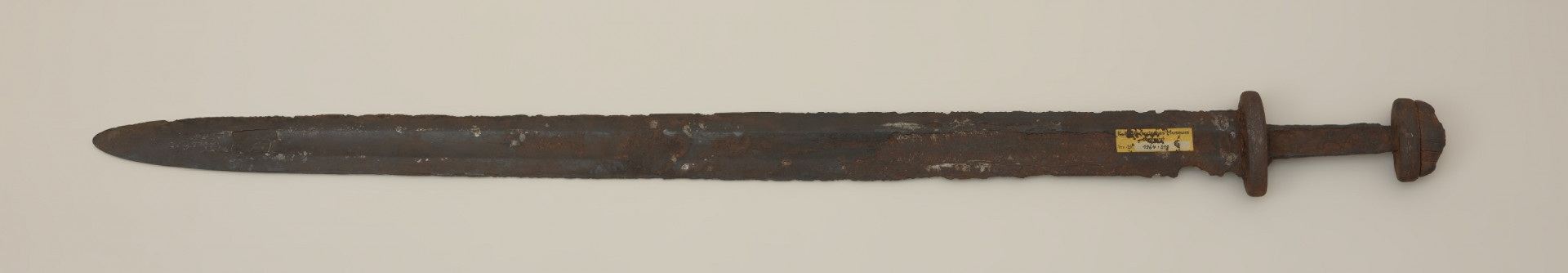miecz dwusieczny - Ujęcie tyłu miecza z góry. Egzemplarz całkowicie zachowany, z korozyjnymi ubytkami. Głownia posiada szerokie zbrocze, widoczne nieomal na całej jej długości. Została wykonana w technice prętowej, metodą dziwerowania. Ślady zasosowania tej techniki widoczne są po obu stronach głowni, z jednej w postaci tzw. „jodełki”, a z drugiej strony jako tzw. „dziwer gwiaździsty”. Świadczy to o użyciu dwóch lub czterech elementów, z których każdy wykonany został z siedmiowarstwowych laminowanych prętów, następnie poddanych skręceniu i finalnie przepołowionych. Następnie dodano nakładki ostrzowe wykonano z warstwowanych taśm o różnym stopniu nawęglania. Rękojeść miecza ma krótki, masywny jelec, który w przekroju bocznym przypomina ośmiobok, oraz dwuczęściową głowicę. Trzpień do rękojeści ma kształt zbliżony do prostokąta i nieznacznie zwęża się ku głowicy. Odkryto na nim ślady organicznych okładzin. Elementy oprawy pierwotnie były ornamentowane w postaci cienkich pionowych pasków wykonanych ze stopu miedzi i cynku.