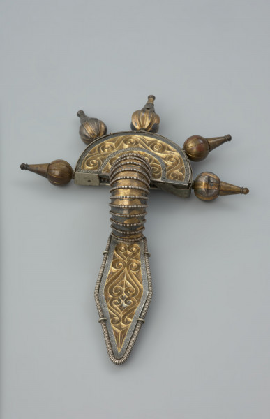 ozdoba; Srebrna, pozłacana zapinka - Ujęcie z tyłu. Srebrna, pozłacana fibula z rombowato ukształtowaną długą nóżką, krótką pochewką i wysoko wysklepionym kabłąkiem. Zapinka ma główkę w postaci półkolistej tarczki nad sprężynką zaopatrzoną w 5 guzów wykonanych ze złotej taśmy.