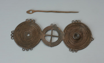 Zapinka trójelementowa - Ujęcie z przodu. Zapinka składa się z dwóch zewnętrznych i jednej centralnej tarczki. Dwie zewętrzne pokrywają koncentryczne żłobki. Na jednej z nich pośrodku znajduje się ozdobny detal w formie guzka. Analogiczny guzek na drugiej tarczce nie zachował się. Na trzeciej, wewnętrznej tarczce o kolistym kształcie wpisany jest krzyż. Na stronie spodniej znajduje się igła będąca elementem zapięcia.