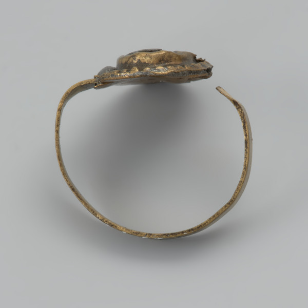 pierścionek z oczkiem - Ujęcie tyłu z góry. Trzyczęściowy brązowy pierścionek z oczkiem, składający się z metalowej taśmy stanowiącej otok, metalowej tarczki pod oczko złożonej z owalnej płaskiej blaszki spodniej oraz dodatkowej taśmy tworzącej eliptyczne gniazdko. Jest ona zdobiona dookoła odciskami. Trzecim elementem, który uzupełnia oczko jest grafitowe szkiełko zachowane jedynie w części, przyklejone do spodniej tarczki i przytrzymywane przez krawędzie otaczającej je taśmy. Między blaszkami oczka pozostawione są dwie 