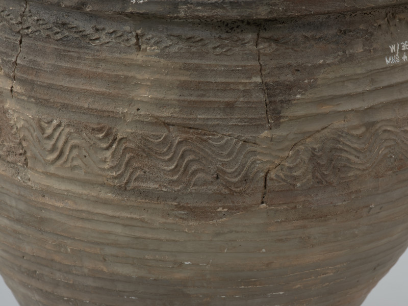 naczynie gliniane - Ujęcie górnego fragmentu tylnej ścianki naczynia. Garnek o baniastej formie z charakterystycznie wychyloną na zewnątrz krawędzią wylewu, obtoczony w całości na kole garncarskim. Brzusiec garnka znajduje się w 2/3 wysokości. Powierzchnia glinianego pojemnika przyozdobiona jest ornamentem wielowątkowym skośnych żłobków, dookolnych linii rytych, linii falistych oraz ponownych linii rytych. Fakturę naczynia tworzy dobrze wyprofilowana, gładka powierzchnia. Garnek wylepiono z glinianych wałeczków z wcześniej przygotowanej masy garncarskiej zawierającej domieszkę drobno i średnioziarnistego tłucznia - piasku i granitu.