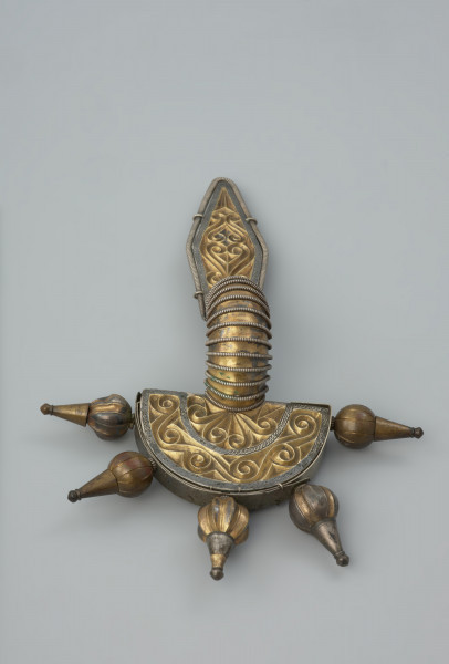 ozdoba; Srebrna, pozłacana zapinka - Ujęcie z przodu. Srebrna, pozłacana fibula z rombowato ukształtowaną długą nóżką, krótką pochewką i wysoko wysklepionym kabłąkiem. Zapinka ma główkę w postaci półkolistej tarczki nad sprężynką zaopatrzoną w 5 guzów wykonanych ze złotej taśmy.