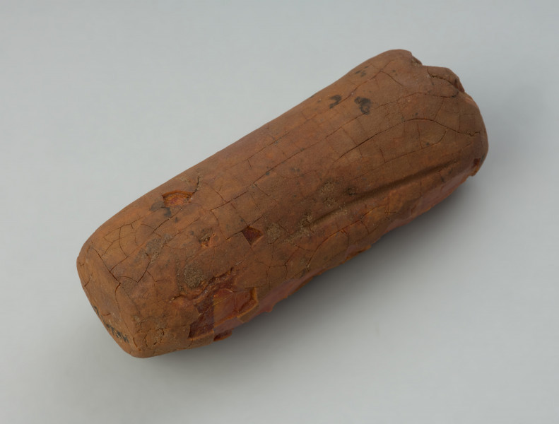 zawieszka cylindryczna; amulet bursztynowy - Ujęcie 3/4 z boku skosem do tyłu. Podłużna, cylindryczna zawieszka wykonana z dużej konkrecji bursztynu, której większość powierzchni pokrywa patyna przypominająca glinę. W szczytowej oraz bocznej ściance znajdują się wywiercone otworki nie przechodzące przez całą grubość przedmiotu.