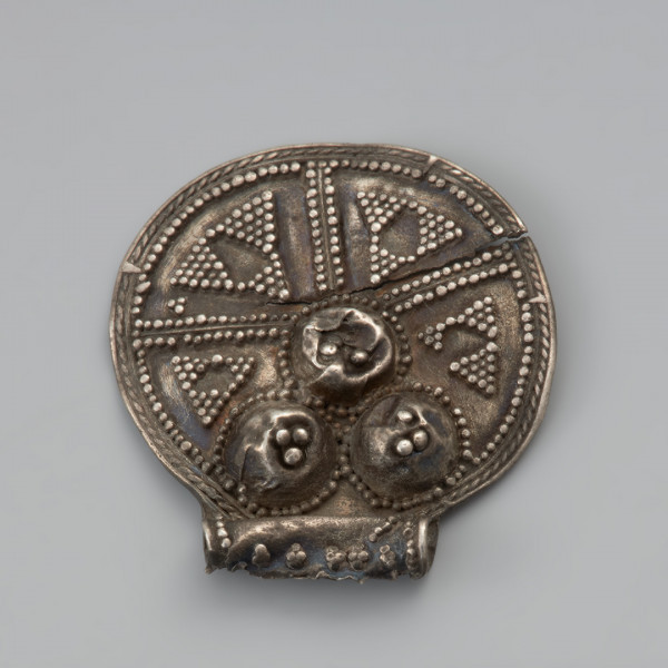 ozdoba; biżuteria; zawieszka - Ujęcie przodu zawieszki uszkiem do dołu. Okrągła zawieszka wycięta ze srebrnej blachy o średnicy 2,5 cm, wraz z pasmem zawiniętym w rurkowate uszko. Zdobiona jest po jednej stronie trzema guzkami, z których każdy jest otoczony wieńcem przylutowanych półkolistych granulek. Również na każdym z guzków znajdują się po trzy granulki. Na pozostałej powierzchni blaszki znajdują się cztery promieniście rozłożone pola wypełnione ornamentem trójkątów wykonanym w technice granulacji oraz łańcuszkiem półjodełki. Uszko, również zdobione jest granulkami. Do tylnej, niezdobionej strony, od boków uszka do około połowy wysokości okrągłej części, przylutowane są wąskie pasemka blachy wzmacniające zawieszkę. Zarówno uszko jak i zawieszka właściwa poniżej linii guzków są pęknięte.