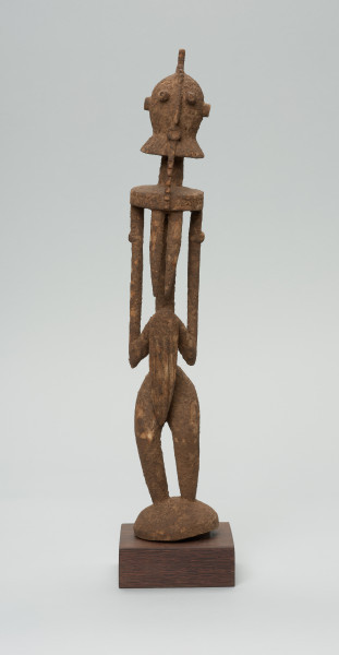 rzeźba - Ujęcie z przodu. Drewniana, rzeźbiona postać znajdująca się w pozycji stojącej, przekrzywiona w lewą stronę. Głowa odcina się od szyi i tułowia prostą kreską. Ramiona proste. Ręce ułożone wzdłuż ciała. Nogi stosunkowo krótkie. Oczy, nos, usta wypukłe, broda w kształcie kryzy.