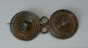 Zapinka binoklowata - Ujęcie z góry przodu zapinki. Zapinka brązowa wykonana z czworograniastego drutu, z którego uformowano dwie spiralne tarczki z ozdobnymi guzami w centrum każdej z nich. Między tarczkami znajduje się dodatkowy, ósemkowato skręcony drut. Na spodniej stronie przytwierdzono centralnie do jednej z tarczek igłę, służącą wpinaniu w odzież. Na drugiej tarczce umieszczono, w jej centralnym punkcie, haczyk (skobelek) do zahaczania igły.