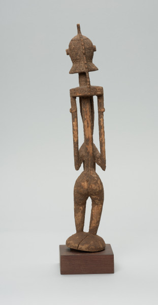 rzeźba - Ujęcie z tyłu. Drewniana, rzeźbiona postać znajdująca się w pozycji stojącej, przekrzywiona w lewą stronę. Głowa odcina się od szyi i tułowia prostą kreską. Ramiona proste. Ręce ułożone wzdłuż ciała. Nogi stosunkowo krótkie. Oczy, nos, usta wypukłe, broda w kształcie kryzy.