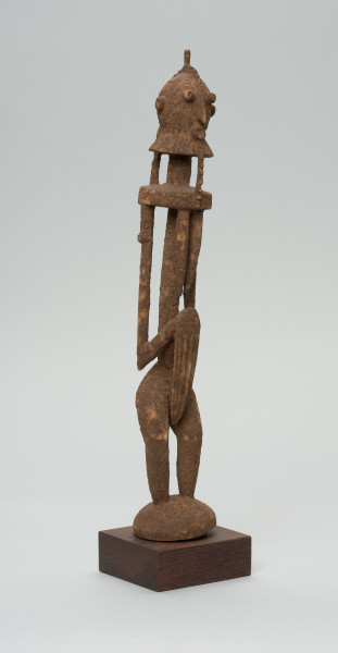 rzeźba - Ujęcie z przodu z prawej strony. Drewniana, rzeźbiona postać znajdująca się w pozycji stojącej, przekrzywiona w lewą stronę. Głowa odcina się od szyi i tułowia prostą kreską. Ramiona proste. Ręce ułożone wzdłuż ciała. Nogi stosunkowo krótkie. Oczy, nos, usta wypukłe, broda w kształcie kryzy.