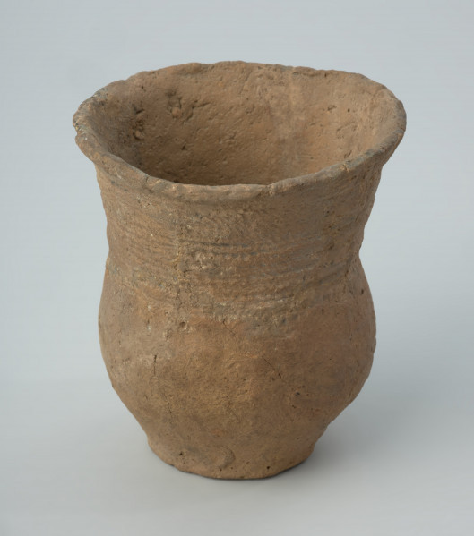 Naczynie gliniane - Ujęcie boku; Puchar o kształcie esowatym, zdobiony na szyjce dookolnymi pasmami odcisków sznura, złożonymi z 4 grup odcisków zwielokrotnionych linii, podkreślonym od dołu dookolną linią drobnych nakłuć. Powierzchnia nierówna, niestarannie wyrównywana, ze śladami po wypadających w trakcie wypału dodatkach w masie garncarskiej. Nad dnem wywiercony wtórnie otworek. Naczynie rekonstruowane i uzupełnione barwionym gipsem.