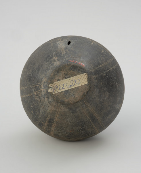 naczynie gliniane - Ujęcie z dołu. Gliniane naczynie wąskootworowe o flaszowatej formie z wysoką cylindryczną szyjką. Powierzchnia jest wyświecana, barwy ciemnobrunatnej. Brzusiec jest dwustożkowaty, zdobiony techniką kółka zębatego motywem meandra schodkowego, a w dolnej partii pionowymi liniami. Szyjkę obiegają u podstawy dwie listy plastyczne.