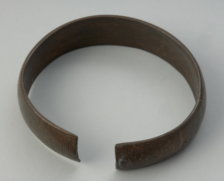 bransoleta mankietowata - Ujęcie z góry. Bransoleta, tworząca szeroką otwartą obręcz, wykonana jest z blachy brązowej. Przez całą długość środkowej części bransolety biegnie wąskie, wysklepione żeberko, które stanowi element dekoracyjny. Po obu jego stronach umieszczono ornament w postaci rytej jodełki, prostokąta i niewielkich łuków.
